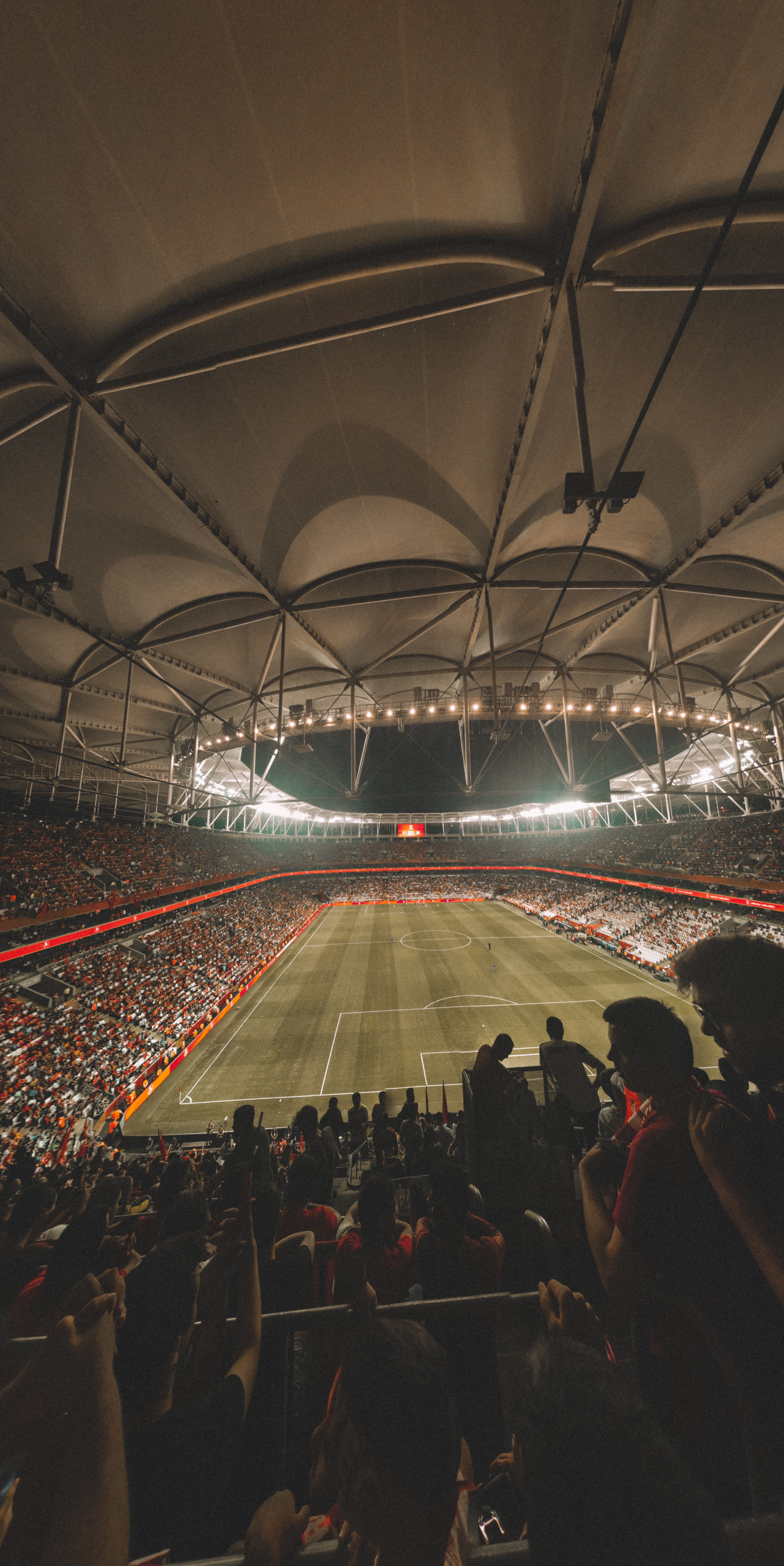 An empty green football pitch under floodlights, seen from above, with spectators in the stands before kick-off